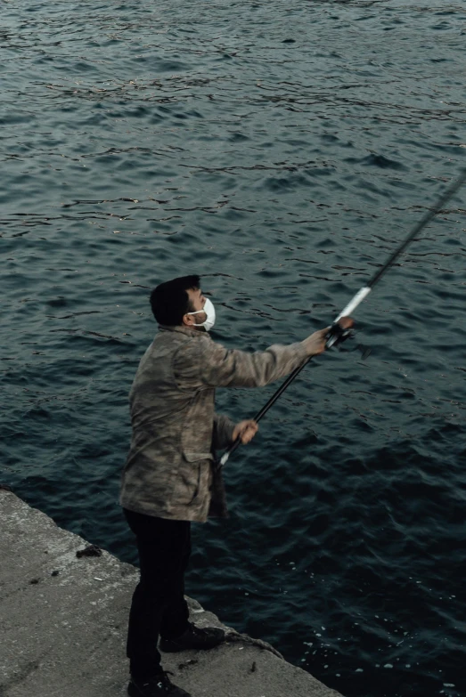 a man standing next to a body of water holding a fishing pole, wearing a mask, lo fi, a high angle shot, grey