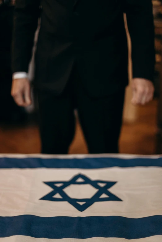 a man in a suit standing next to a flag, by Niko Henrichon, symbolism, casket, israel, on a wooden desk, square