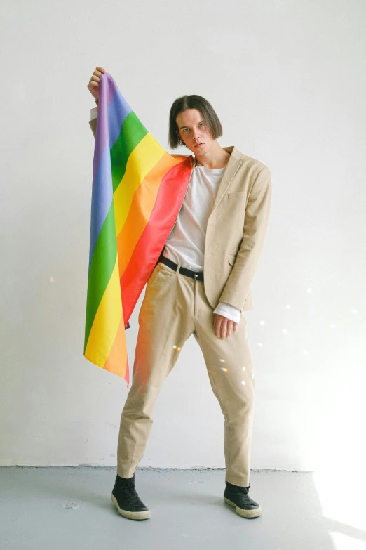 a man holding a rainbow flag in front of a white wall, an album cover, by Nina Hamnett, unsplash, tan suit, jamie campbell bower, wearing pants, holding a white flag