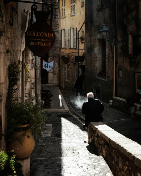 a man sitting on a ledge in a narrow street, inspired by Jean Tabaud, pexels contest winner, renaissance, on a village, ray of light through white hair, france, smoking outside