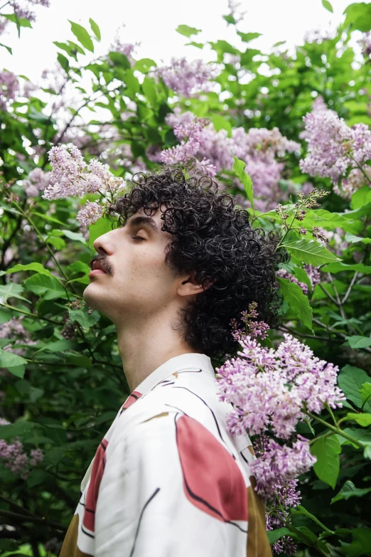 a man standing in front of a bush of purple flowers, an album cover, trending on pexels, curly hair, enes dirig, lily frank, delicate androgynous prince