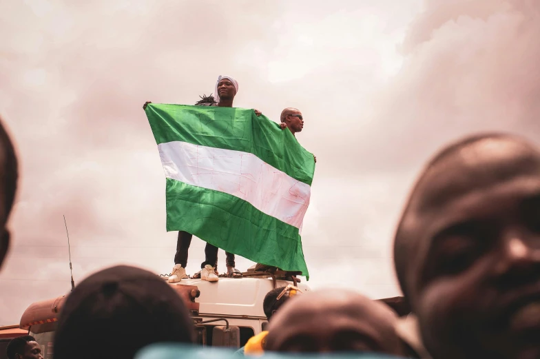 two men standing on top of a truck holding a flag, a photo, by Matija Jama, trending on pexels, skin painted with green, crowd of people, obunga, 🚿🗝📝