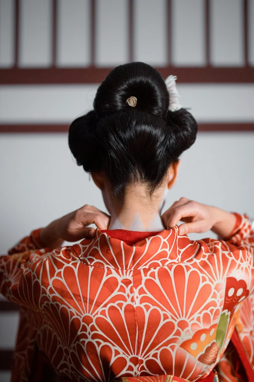 a woman wearing a red and white kimono, hair styled in a bun, showing her shoulder from back, hands in her hair