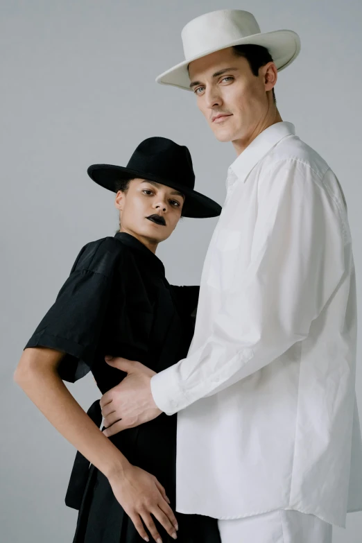 a man and a woman standing next to each other, an album cover, unsplash, bauhaus, white hat, fashion studio lighting, pose 4 of 1 6, wearing modern gothic clothes