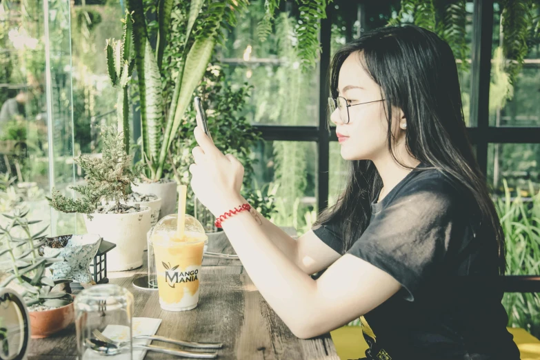 a woman sitting at a table with a glass of orange juice, pexels contest winner, realism, asian female, next to a plant, profile image, selfie shot straight on angle