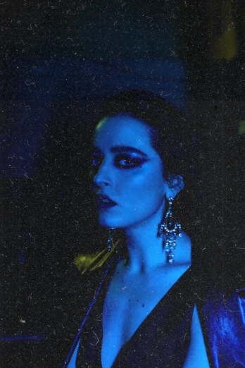 a woman in a black dress standing in a dark room, an album cover, inspired by Elsa Bleda, unsplash, bauhaus, glowing blue face, huge earrings and queer make up, low quality grainy, nighttime scene