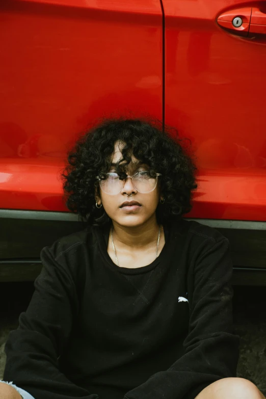 a woman sitting on the ground next to a red car, an album cover, pexels contest winner, jayison devadas, wearing black frame glasses, ((portrait)), curly haired