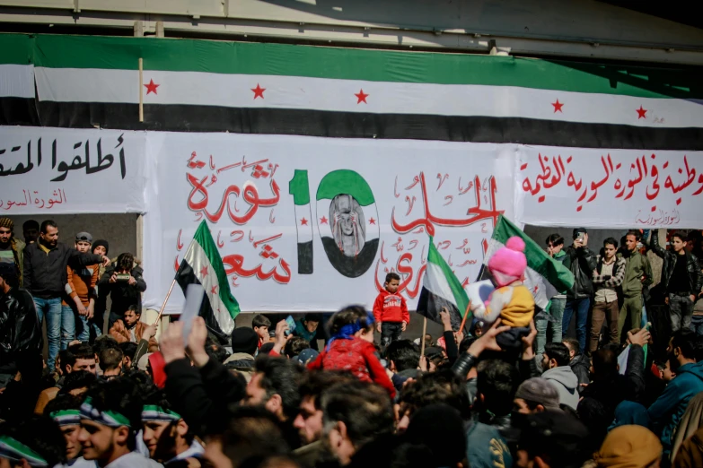 a large group of people standing in front of a banner, by Bouchta El Hayani, shutterstock, hurufiyya, square, real life photo of a syrian man, winning, february)