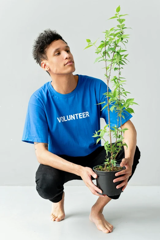 a man kneeling down holding a potted plant, an album cover, inspired by John Luke, visual art, blue tight tshirt, wearing a marijuana t - shirt, profile image, vp of marketing