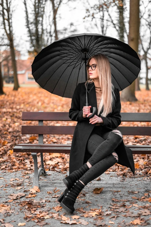 a woman sitting on a bench holding an umbrella, by Zofia Stryjenska, pexels contest winner, black outfit, wearing black frame glasses, panoramic view of girl, autumnal