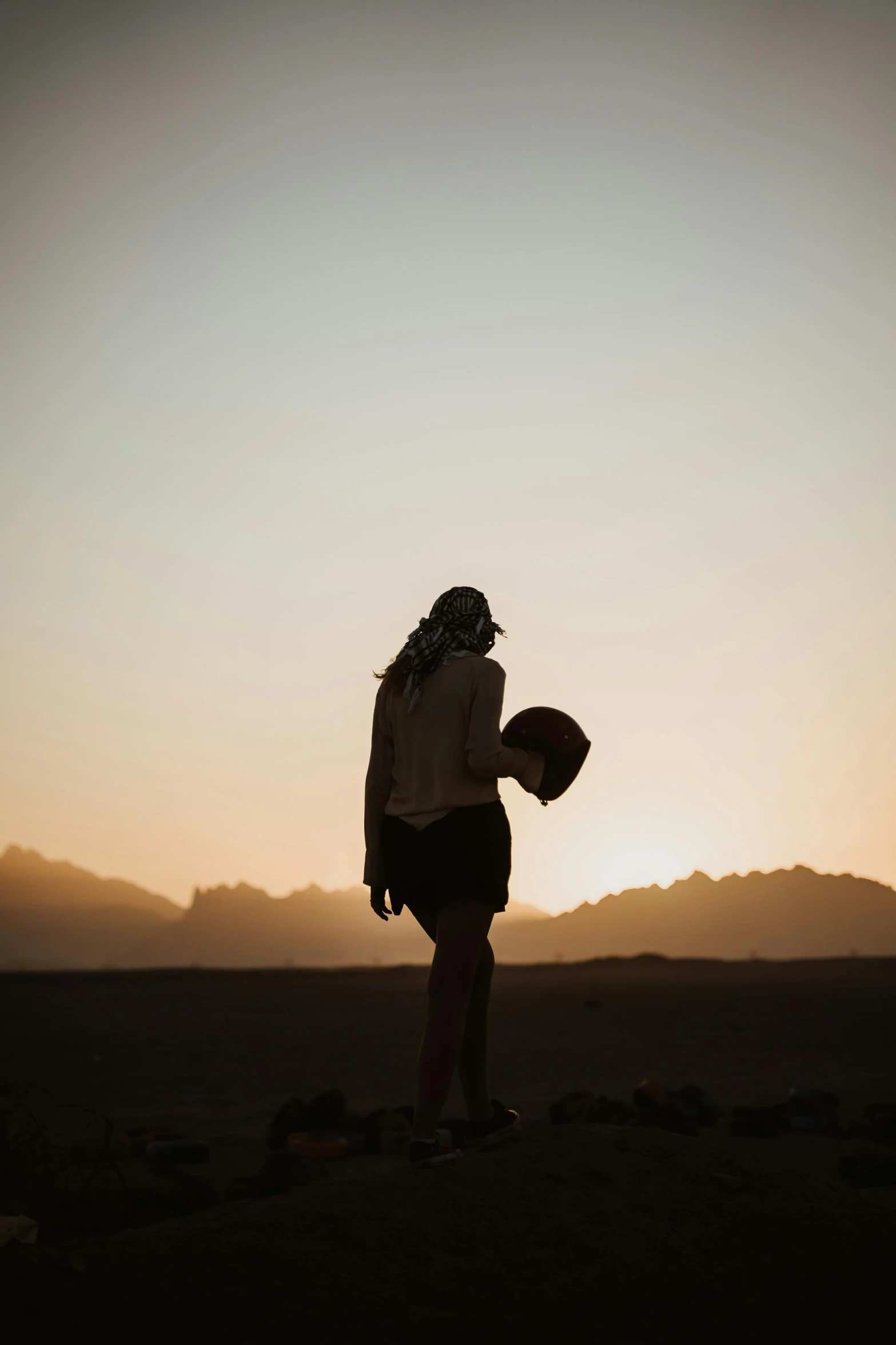 a person standing in the middle of a desert, holding helmet, ((sunset)), woman, trending photo