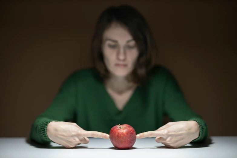 a woman sitting at a table with an apple in front of her, inspired by Anna Füssli, unsplash, hyperrealism, shrugging arms, uneven glass apple in the dark, person in foreground, holding it out to the camera