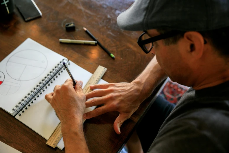 a man sitting at a table writing on a notebook, a drawing, by Joe Stefanelli, process art, fan favorite, quality draughtmanship, a high angle shot, crafts