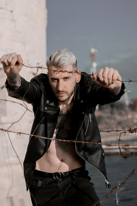 a man in a leather jacket leaning against a fence, an album cover, inspired by Elsa Bleda, trending on pexels, beefcake pose, silver hair, barbed wire, hasan piker