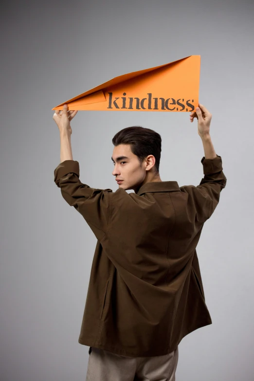 a man holding a sign that says kindness, an album cover, inspired by Alexander Kanoldt, brown ) ), asian man, grey orange, promo photo