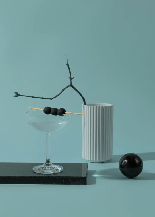 a martini sitting on top of a table next to a vase, inspired by Carlo Martini, flying black marble balls, black vertical slatted timber, porcelain organic, black and teal paper