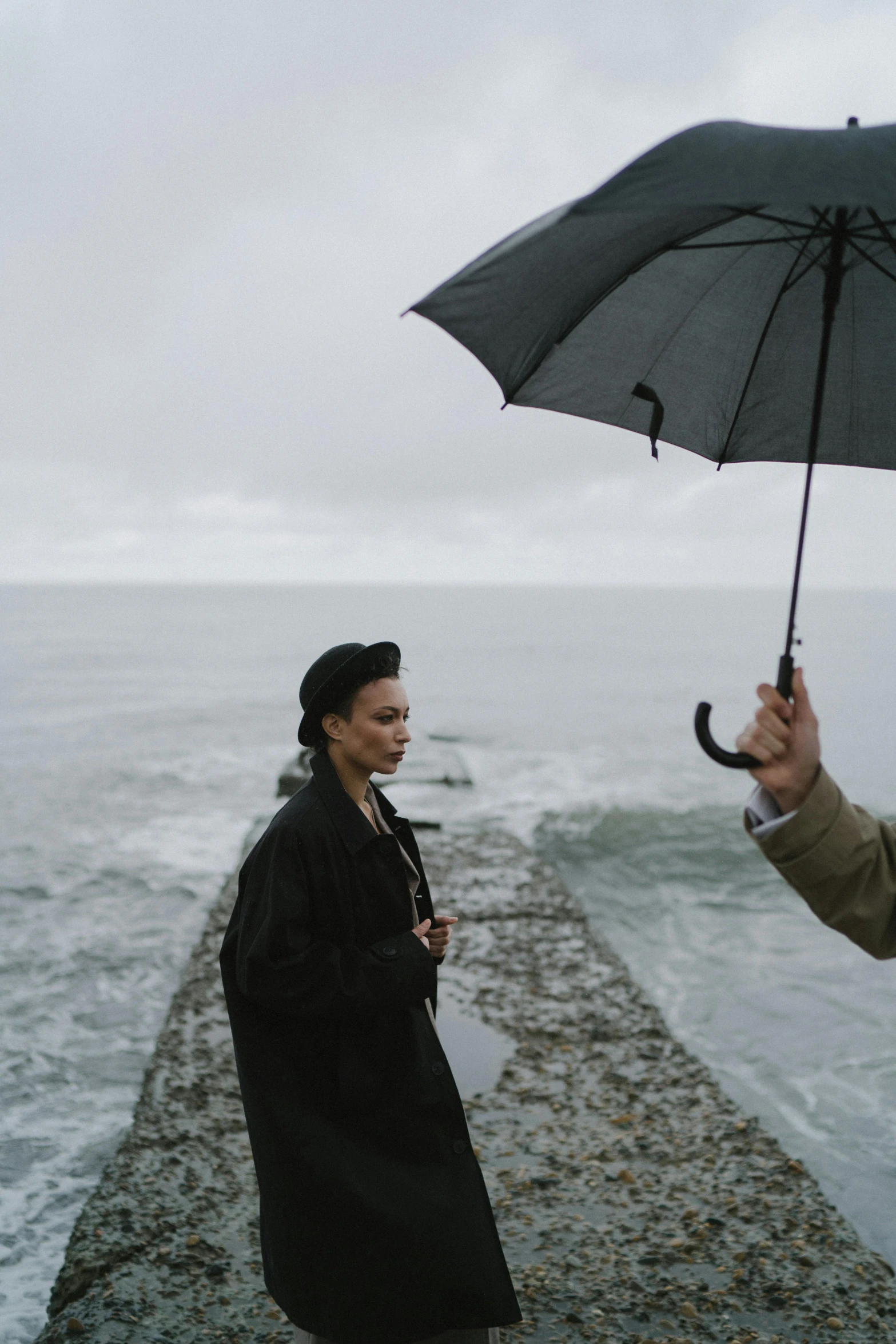 a man standing next to a woman holding an umbrella, inspired by Peter Lindbergh, unsplash, visual art, on the coast, b - roll, on set, still from a live action movie