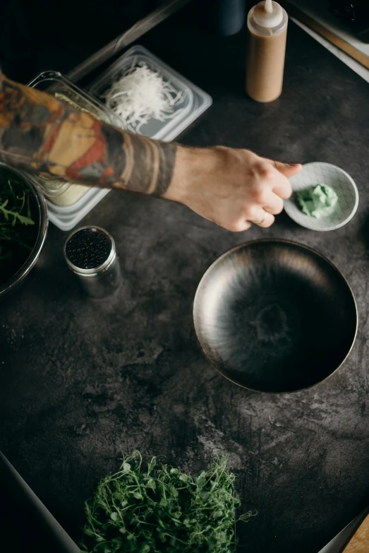 a close up of a person preparing food on a table, a still life, inspired by Nishida Shun'ei, trending on unsplash, dark matte metal, herbs, bowl, tattooed