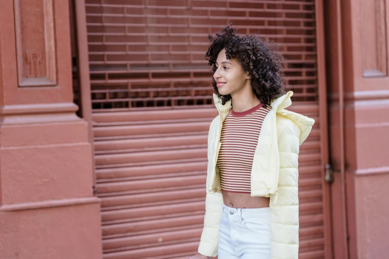 a woman with curly hair standing in front of a building, trending on pexels, puffer jacket, faded red and yelow, striped, white jacket