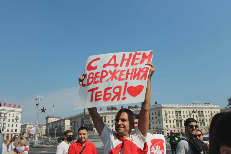 a man holding a sign in the middle of a crowd, a poster, socialist realism, red hearts, neo norilsk, on a bright day, saying