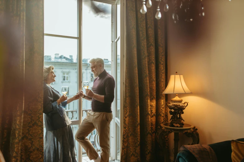 a man and a woman standing in front of a window, by Emma Andijewska, pexels contest winner, renaissance, themed after wine, hotel room, two old people, directoire style