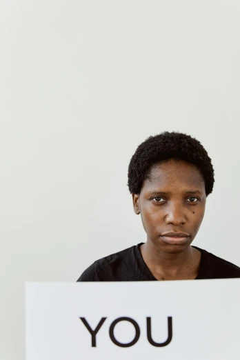 a man holding a sign in front of a white wall, an album cover, by Lily Delissa Joseph, trending on unsplash, computer art, african woman, looking at monitor, serious facial expression, sitting in front of computer