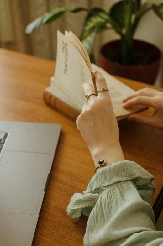 a woman reading a book in front of a laptop, by Julia Pishtar, trending on pexels, wearing two silver bracelets, dusty library, japanese collection product, point finger with ring on it