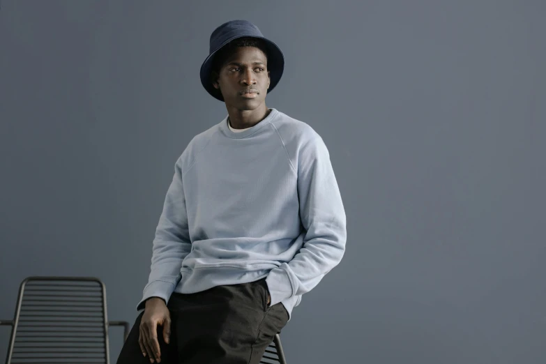 a man sitting on a chair wearing a hat, a character portrait, inspired by Theo Constanté, trending on unsplash, wearing sweatshirt, pale blue, confident relaxed pose, lance reddick