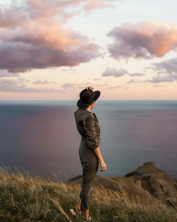 a woman standing on top of a hill next to the ocean, pink skies, wearing a straw hat and overalls, new zeeland, trending photo