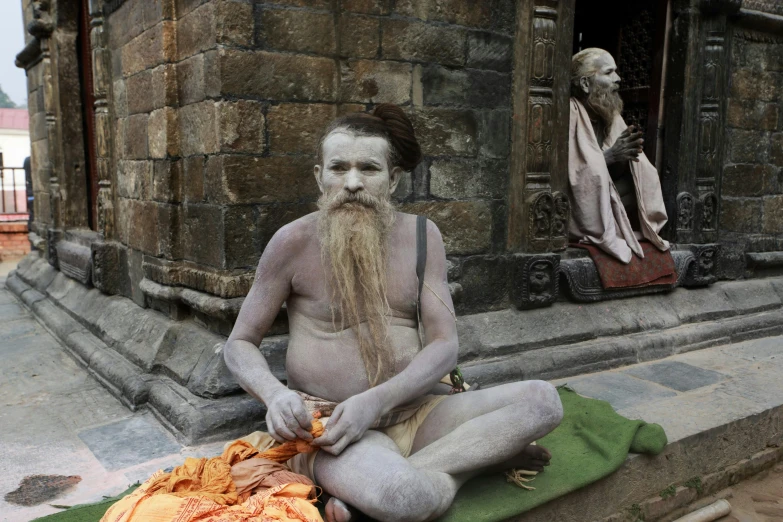 a man sitting on the ground in front of a building, a statue, reddit, bengal school of art, hairy orange skin, dressed as an oracle, loincloth, grey mustache