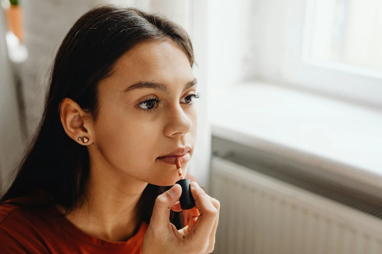 a woman putting on lipstick in front of a window, trending on pexels, antipodeans, portrait sophie mudd, with brown skin, manuka, with a pointed chin