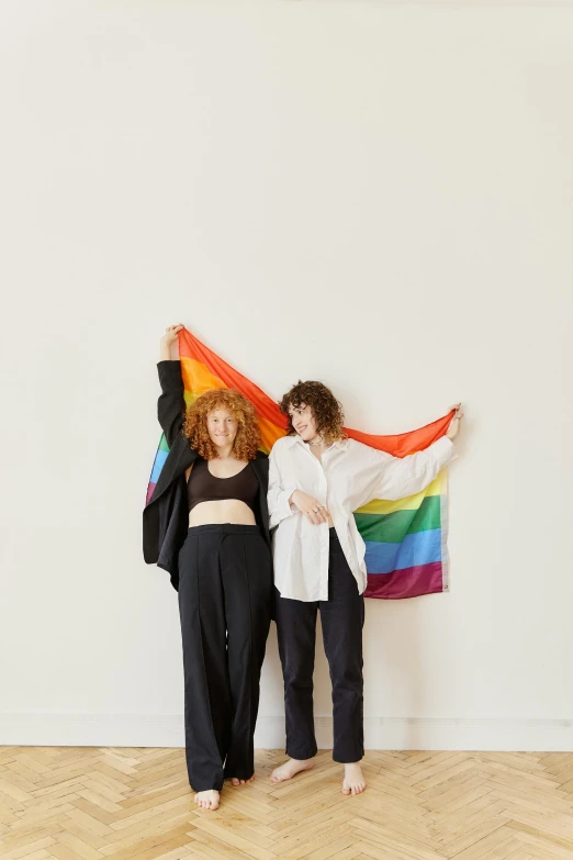 two women standing next to each other holding a rainbow flag, an album cover, unsplash, antipodeans, brunette boy and redhead boy, in a white room, curly haired, flag in hands up