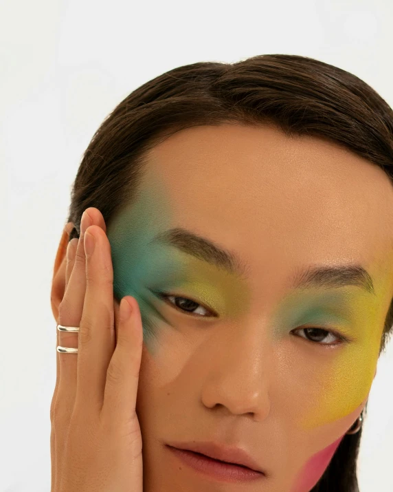 a woman with colorful makeup holding her hand to her face, inspired by Russell Dongjun Lu, androgyny, zoomed out shot, showing forehead, asian male