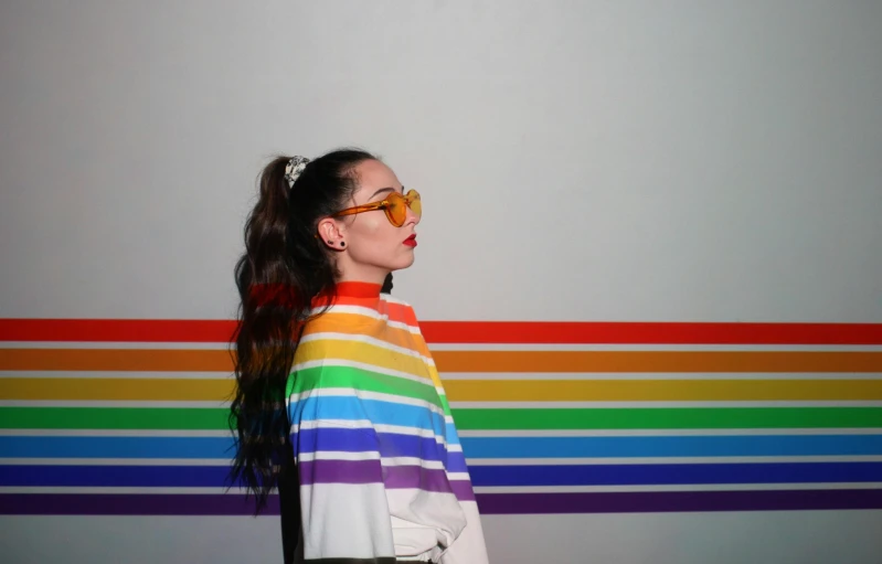 a woman standing in front of a rainbow striped wall, an album cover, inspired by Julia Pishtar, trending on pexels, with glasses, sasha grey, bisexual lighting, profile image