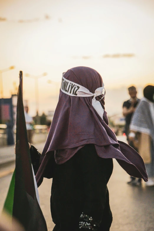 a woman walking down a street holding a flag, by Maryam Hashemi, trending on unsplash, hurufiyya, wearing a headband, protest, sundown, maroon and white