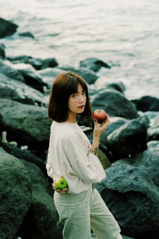 a woman standing on a rocky beach holding an apple, inspired by Yuko Tatsushima, ulzzang, with a bob cut, leng jun, **cinematic