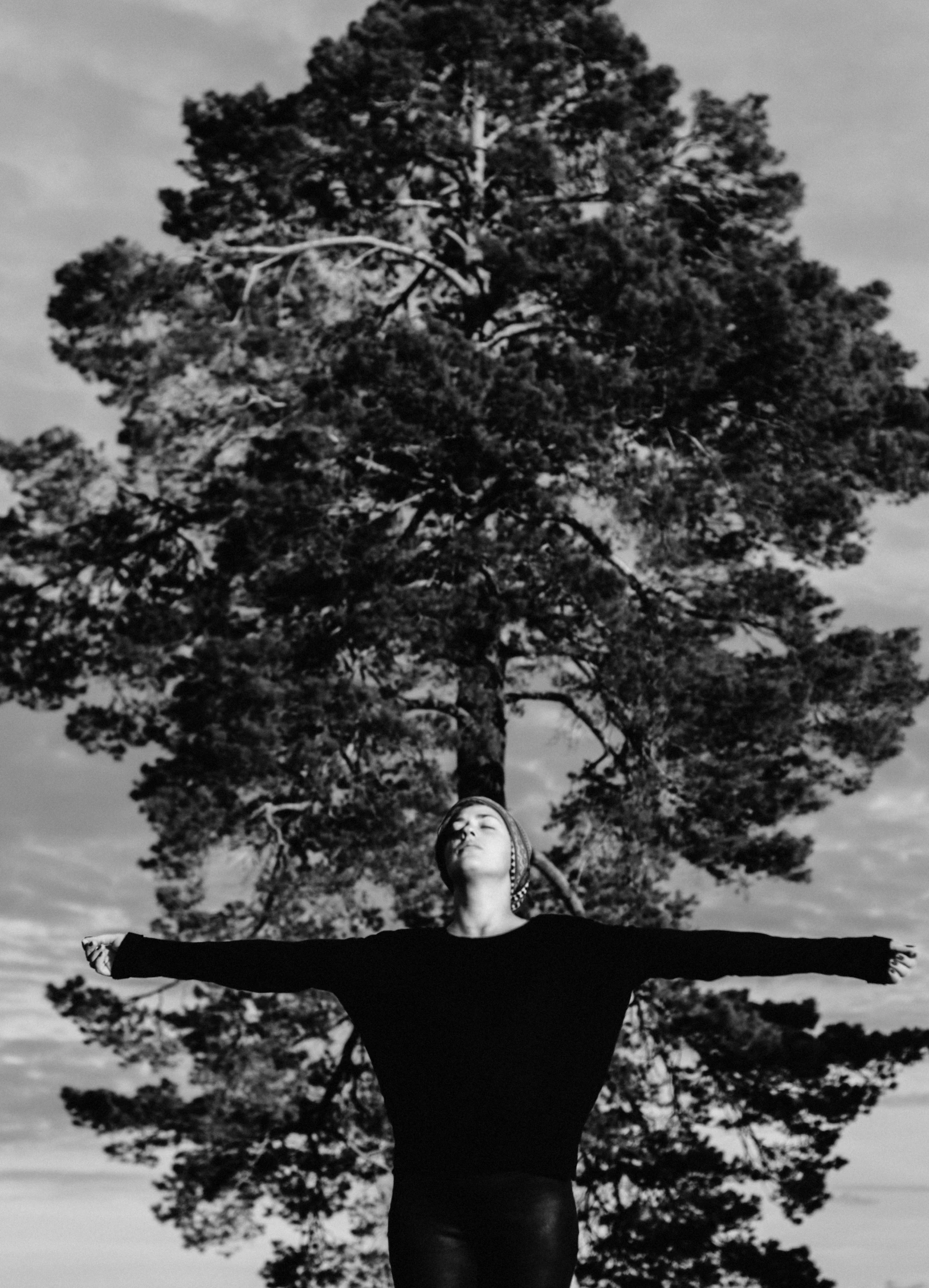 a black and white photo of a woman standing in front of a tree, a black and white photo, by Emma Andijewska, unsplash, symbolism, his arms spread. ready to fly, with matsu pine trees, yung lean, displayed