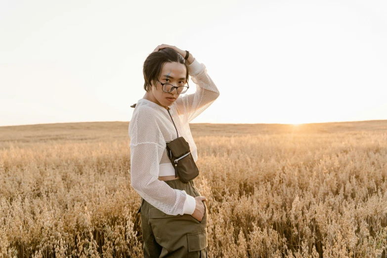 a woman standing in a field of tall grass, inspired by Ren Hang, trending on pexels, wearing square glasses, holding a gold bag, androgynous male, wearing cargo pants