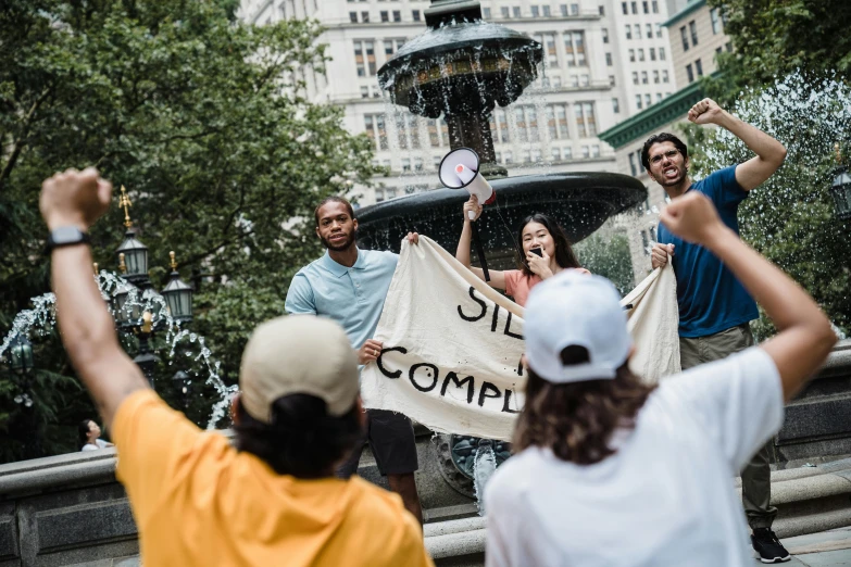 a group of people holding a sign in front of a fountain, by Nina Hamnett, unsplash contest winner, sisyphus compostition, attacking nyc, sza, i és complex