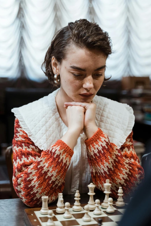 a woman sitting at a table playing a game of chess, wearing a cardigan, daisy ridley, medium head to shoulder shot, student