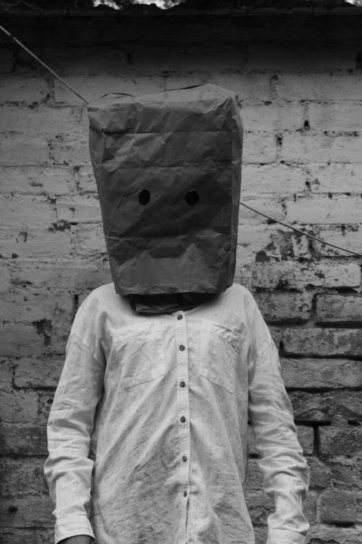 a man standing in front of a brick wall with a paper bag on his head, an album cover, inspired by Roger Ballen, unsplash, anthropomorphic _ humanoid, square face, monochrome, woman