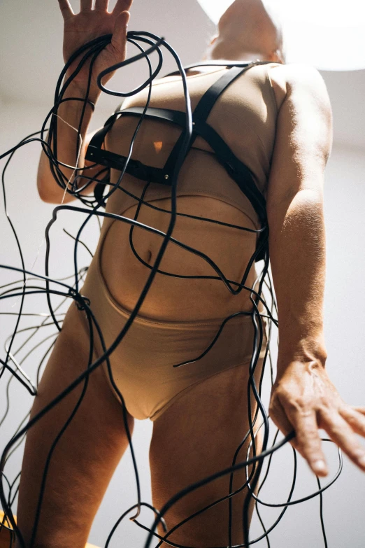 a man standing on top of a bed covered in wires, an abstract sculpture, inspired by Hans Bellmer, unsplash, photograph of a techwear woman, wearing leotard, revealing wires and electronics, tan