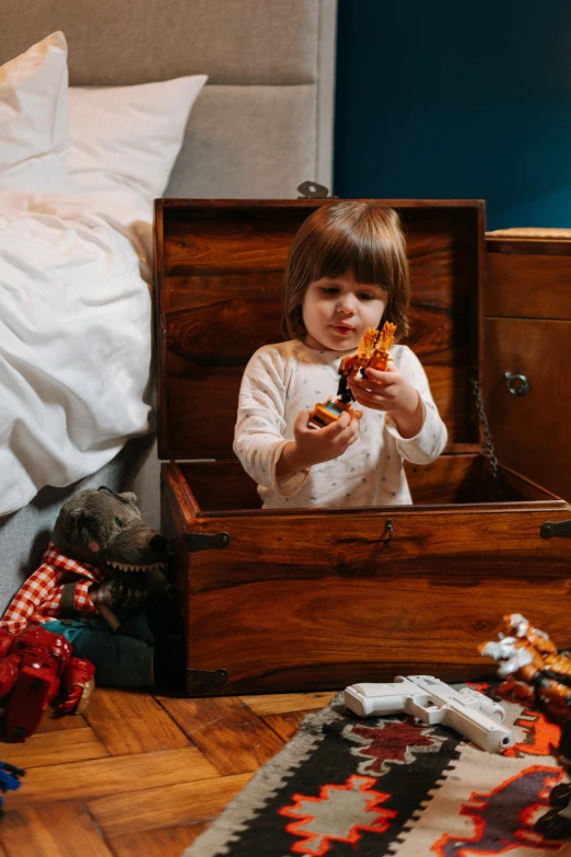 a little girl that is sitting in a trunk, looking at the treasure box, in a bedroom, holding a wood piece, snacks