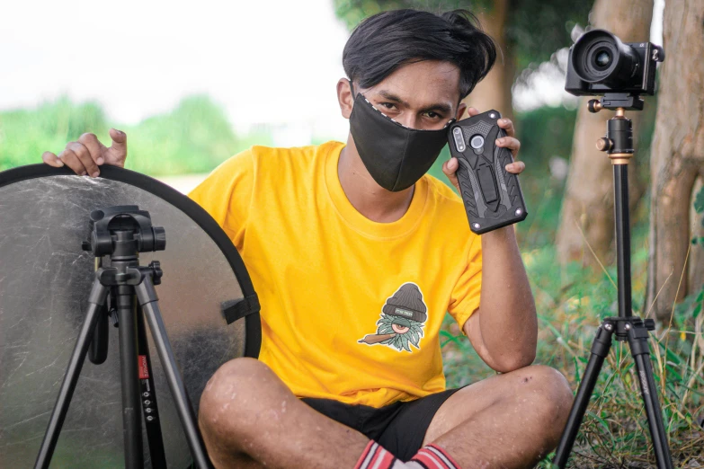 a man sitting on the ground holding a camera, inspired by Rudy Siswanto, wearing all black mempo mask, wearing a modern yellow tshirt, film cam, wearing bandit mask