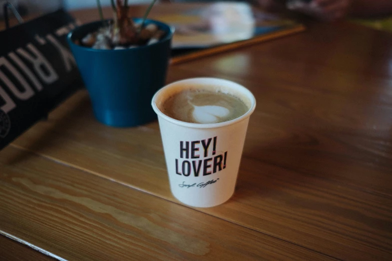 a cup of coffee sitting on top of a wooden table, hey buddy, hotoverse, soymilk, high quality product image”