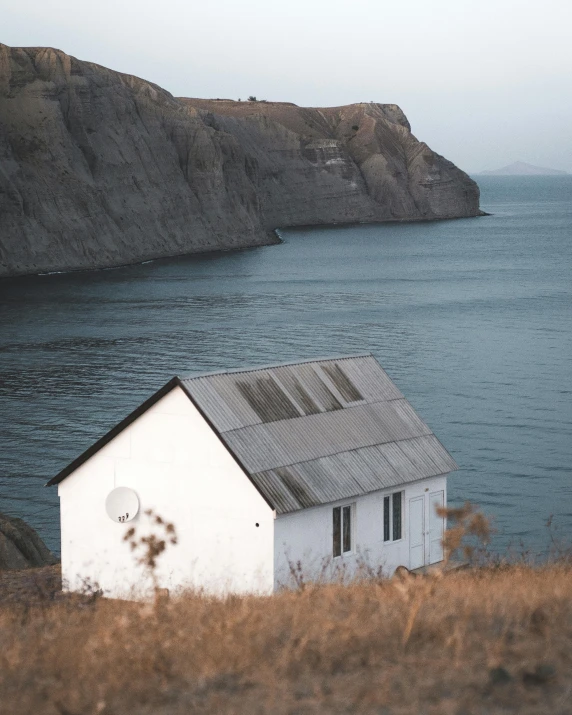 a small white house sitting on top of a hill next to a body of water, pexels contest winner, black sea, slate, shed, casually dressed