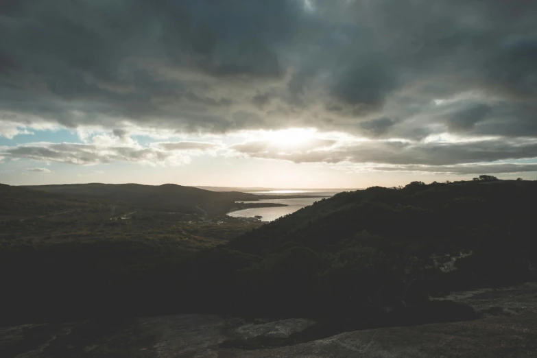 a large body of water sitting on top of a lush green hillside, unsplash contest winner, australian tonalism, moody sunset and dramatic sky, looking down on the view, grey, low quality footage