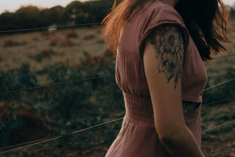 a woman standing in a field next to a fence, a tattoo, pexels contest winner, symbolism, faded pink, shoulder tattoo, still from a movie, portrait featured on unsplash