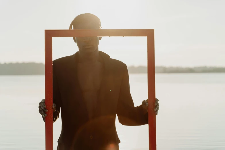 a person holding a picture frame in front of a body of water, an album cover, pexels contest winner, surrealism, man is with black skin, wearing a black and red suit, backlight body, ignant