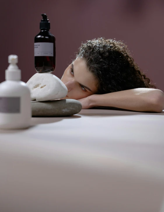 a woman laying on top of a table next to a bottle of liquid, healing pods, pouty, wearing a grey robe, profile image